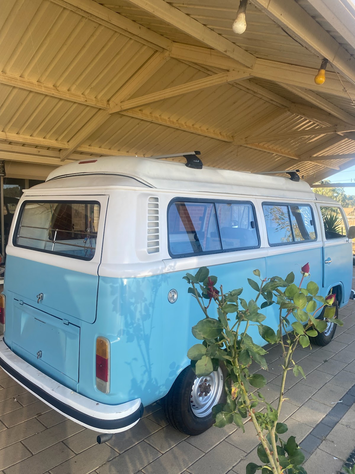 A pale blue and white VW Kombi parked under a carport with its windows fitted